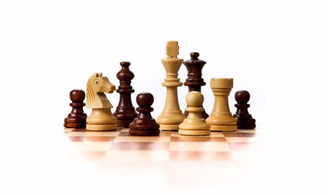 DB Endgame 2026 A small group of light and dark wooden chess pieces, including a king, queen, knight, rook, and pawns, arranged closely together on a chessboard against a white background.